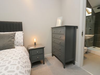 A bedroom with a bed side table with a lit lamp and a chest of drawers next to an open door to a bathroom at Brimstone Backbarrow near Ulverston
