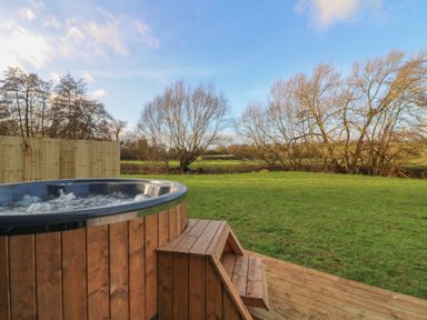 An outdoor hot tub with wooden panels and steps on a wooden deck overlooking a grassy field with bare trees at Squirrel's Nest in South Luffenham near Edith Weston
