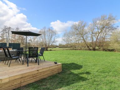 An outdoor wooden deck with a table umbrella chairs and lawn surrounded by trees at Squirrel's Nest in South Luffenham near Edith Weston