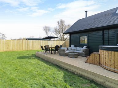 A wooden deck with outdoor seating furniture and a hot tub next to a dark exterior wall of a house in a fenced yard at Squirrel's Nest in South Luffenham near Edith Weston