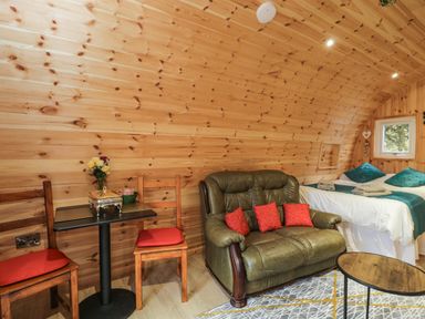 A small room with wood paneled walls and ceiling containing a green leather loveseat with red cushions a bed with teal pillows and towels two wooden chairs with red cushions and a small black table with flowers and a decorative box at The Poddy in Bushton near Lyneham
