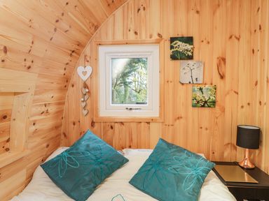 A bedroom with wooden walls and ceiling a bed with two blue pillows a small window floral wall art and a nightstand with a lamp at The Poddy in Bushton near Lyneham
