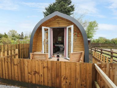 A wooden pod with open doors and a small patio table with chairs and wine glasses at The Poddy in Bushton near Lyneham