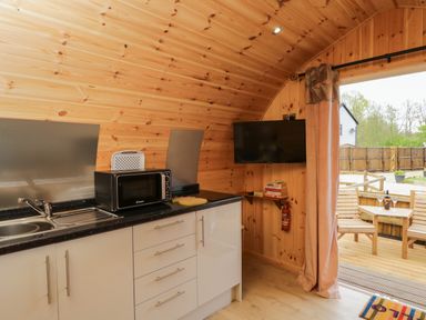 A kitchen area with a microwave on the counter a wall-mounted tv and an open door showing a wooden deck at The Poddy in Bushton near Lyneham