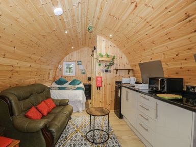 A wooden cabin interior with a bed green sofa small round table and kitchenette at The Poddy in Bushton near Lyneham