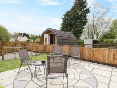 An outdoor patio area with four metal chairs and two small tables near a wooden pod and fenced garden at The Poddy in Bushton near Lyneham