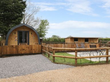 A wooden pod with a fenced patio and outdoor chairs next to a wooden stable at The Poddy in Bushton near Lyneham