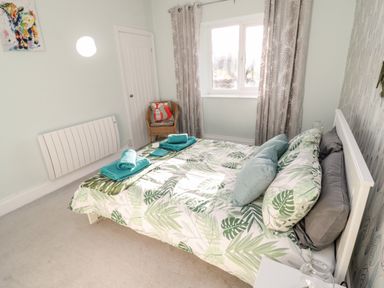 A bedroom with a double bed with leaf-patterned bedding and rolled towels on it a window with curtains a wicker chair with cushions and a door at Moorlaws Farm in Alnwick