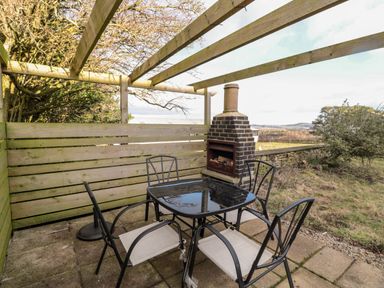 An outdoor patio with glass top table four metal chairs wooden fence brick fireplace and grassy area at Moorlaws Farm in Alnwick