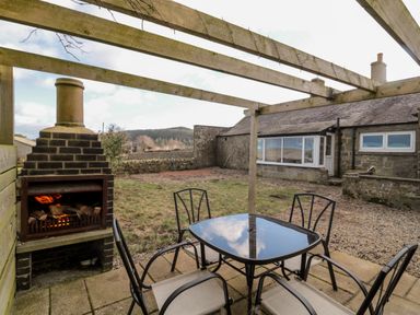 An outdoor patio with a glass table and four chairs under a wooden pergola next to a lit brick barbecue at Moorlaws Farm in Alnwick