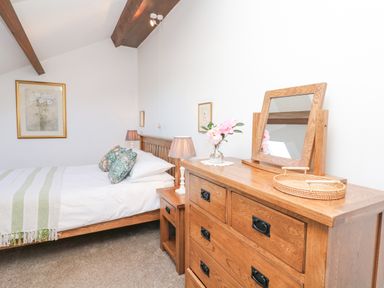 A bedroom with a wooden bed with pillows and a blanket a wooden dresser with a mirror and a vase of flowers at Woodland View in Muddiford near West Down