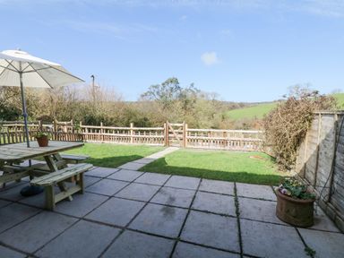 A garden patio with a wooden picnic table umbrella and flowerpot with grass and wooden fencing at Woodland View in Muddiford near West Down