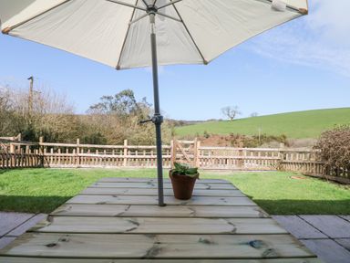A wooden picnic table with a potted plant and a large white umbrella in a garden with a wooden fence and green fields at Woodland View in Muddiford near West Down