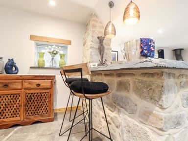 A kitchen bar with high stools and stone counter beside a wooden cabinet with decorative items at La Petit Maison in St Columb Major