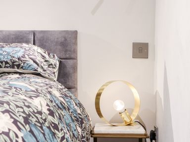 A bedroom corner with a floral bedspread a padded headboard a modern circular lamp on a bedside table at La Petit Maison in St Columb Major