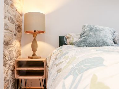 A bedside table with a lamp next to a bed with pillows and patterned bedding at La Petit Maison in St Columb Major