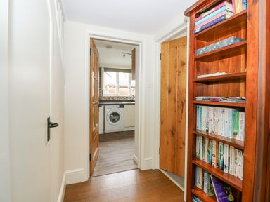 A hallway with a wooden bookshelf filled with books and an open door leading to a kitchen with a washing machine at Easter Cottage in Dersingham