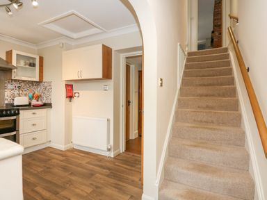 A kitchen area with stove and cabinets next to carpeted stairs and a hallway at Easter Cottage in Dersingham