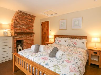 A bedroom with a wooden bed and floral bedding two bedside tables with lamps a brick fireplace and a white chest of drawers at Easter Cottage in Dersingham