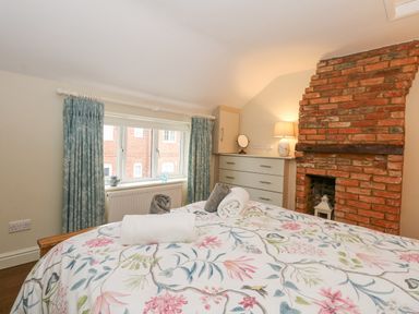 A bedroom with a bed with floral bedspread towels on the bed a chest of drawers exposed brick fireplace and window with blue curtains at Easter Cottage in Dersingham
