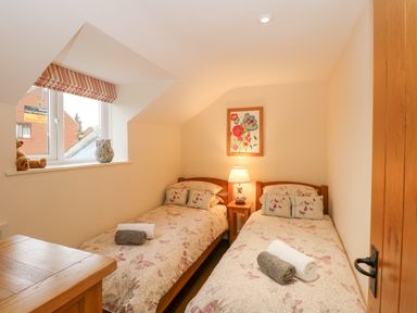 A small bedroom with two single beds with butterfly bedspreads a wooden nightstand with a lamp a window with stuffed animals and a framed floral print on the wall at Easter Cottage in Dersingham