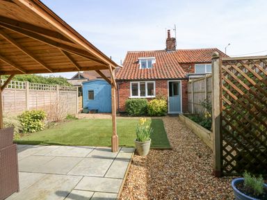 A backyard garden with a small brick house a blue door a blue shed a wooden pergola and gravel paths at Easter Cottage in Dersingham