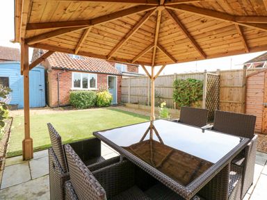 A garden patio with a wooden gazebo covering a glass table and wicker chairs at Easter Cottage in Dersingham