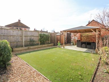 A backyard with a lawn gravel pathway wooden fence raised flower beds and a wooden gazebo with outdoor furniture at Easter Cottage in Dersingham