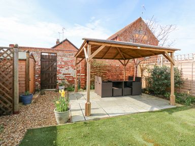 A garden with a wooden pergola covering outdoor seating on a paved area and plants in pots at Easter Cottage in Dersingham