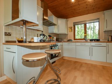 A kitchen with light wood countertops white cabinets stools and a window showing trees at 4 The Glade in St Minver