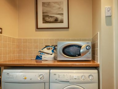 A laundry room with a washing machine, dryer, microwave, and iron on a wooden countertop at 4 The Glade in St Minver