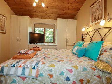 A bedroom with a double bed with patterned bedding and pillows white wardrobes a small television on a dresser under a window and wooden ceiling at 4 The Glade in St Minver