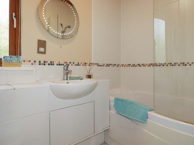 A bathroom with a sink, round mirror, bathtub with a blue towel, and mosaic tile border at 4 The Glade in St Minver
