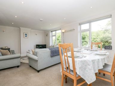 A living room with sofas a dining table set with bowls and glasses and large windows showing trees at Godrevy in Porthcurno