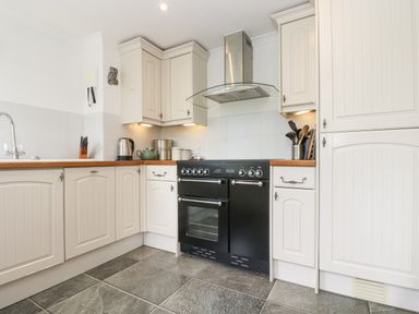 A kitchen with cream cabinets a black stove and oven a stainless steel range hood and tiled floor at Godrevy in Porthcurno