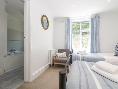 A bedroom with two single beds a window with blue curtains and an armchair at Godrevy in Porthcurno