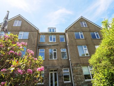 The front view of a multi-story building with windows surrounded by pink flowers and green bushes at Godrevy in Porthcurno