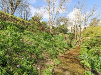 A garden path surrounded by flowering plants and trees with a house in the background at Godrevy in Porthcurno