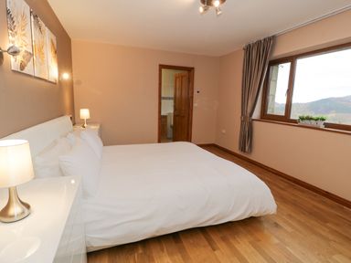 A bedroom with a large bed white bedding bedside tables lamps a window with curtains and a door to an en suite bathroom at Y Tylwyth in Blaenau Ffestiniog