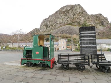 A small green train engine and two black rail carts on a paved area in front of a sign with a rocky hill and houses in the background at Y Tylwyth in Blaenau Ffestiniog