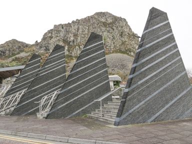 A series of triangular stone structures with horizontal metal strips along a pathway with stairs and a rocky hill in the background at Y Tylwyth in Blaenau Ffestiniog