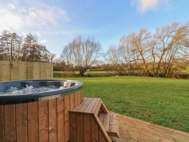A wooden hot tub with steps on a deck in a grassy yard with leafless trees and a wooden fence at Otter's View in South Luffenham near Edith Weston