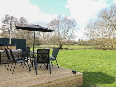 A wooden deck with a glass table umbrella and six chairs overlooking a grassy field with trees at Otter's View in South Luffenham near Edith Weston