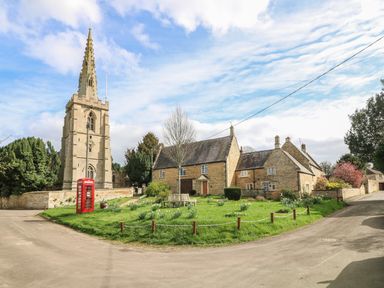 A stone church with a tall spire next to a red telephone box and stone houses with a grassy area and benches at Otter's View in South Luffenham near Edith Weston