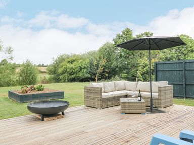 A patio with a rattan sectional sofa, a glass-top table, umbrella, fire pit, raised garden bed, and grass lawn at Otter's View in South Luffenham near Edith Weston