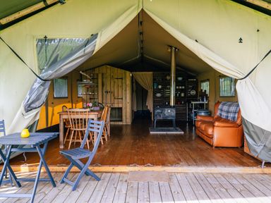 An open tent with wooden flooring containing dining table with chairs a wood stove a leather sofa and shelves with kitchen items at Oak Stockland near Dalwood