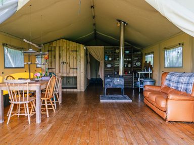 An interior of a tent with a wooden dining table and chairs a wood stove a leather sofa and a wooden kitchen area at Beech in Dalwood