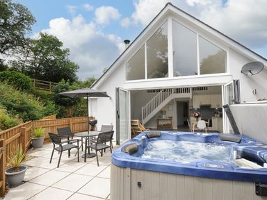 An outdoor patio area with a hot tub table and chairs and an umbrella outside a house with large windows and open doors at Otters Leap in Bideford