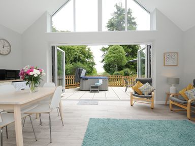 A living area with dining table with flowers and chairs and open doors leading to a patio with hot tub and garden at Otters Leap in Bideford