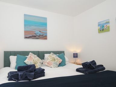 A bedroom with a bed with blue and patterned cushions two piles of folded towels a nightstand with a blue lamp and paintings on the walls at Otters Leap in Bideford